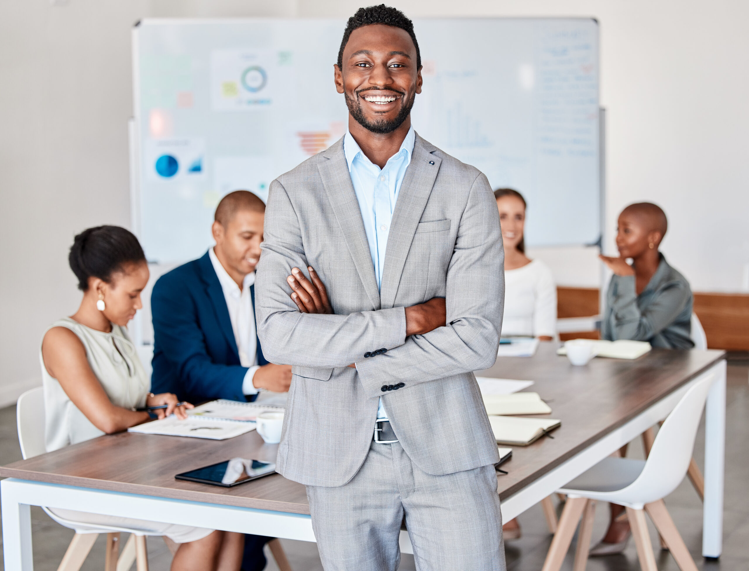 A young business professional smiling and standing with his arms crossed in the middle of a boardroom.